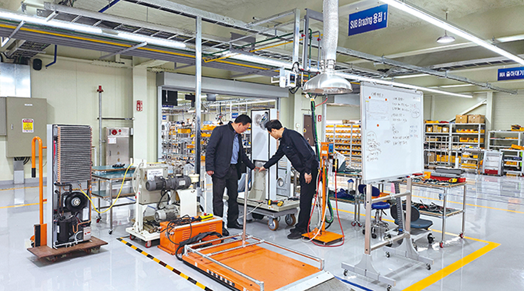 Two workers inspecting automated equipment on the manufacturing floor of a smart factory. Various machines, a whiteboard, and piping systems are arranged together.